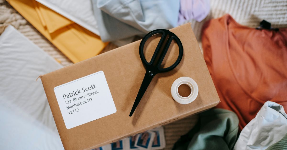Top view of tape with scissors on carton box with exact address and name among clothes